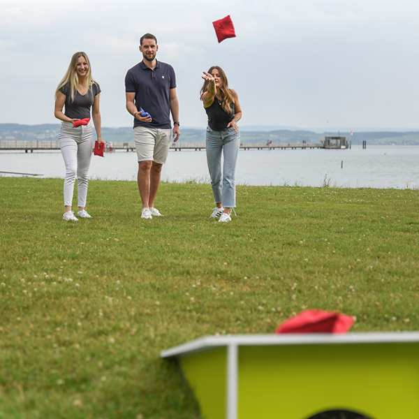Drei Personen beim Cornhole-Spiel auf der Wiese am See