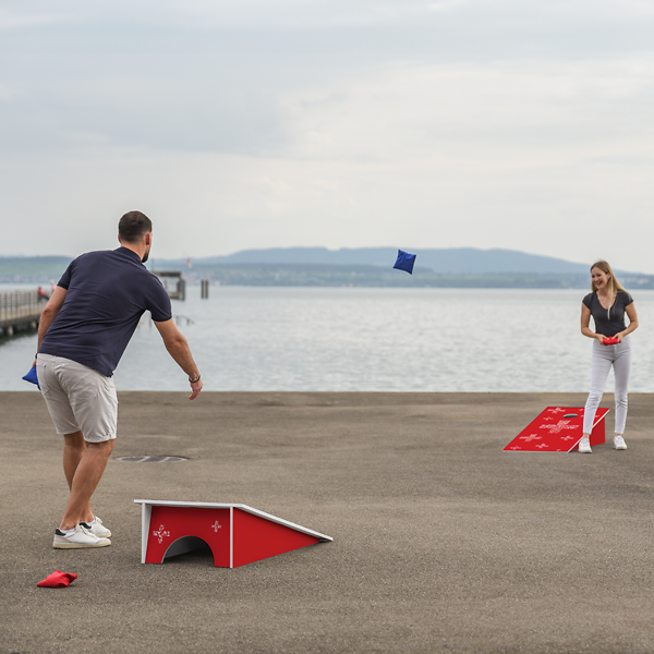 Zwei Personen spielen Cornhole mit roten Brettern am See