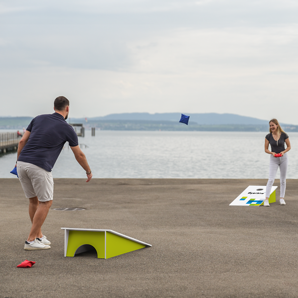 Zwei Personen spielen Cornhole mit bedruckten Brettern am See